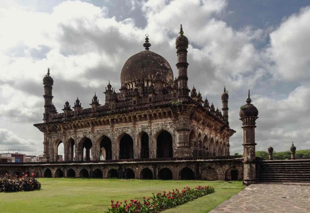 The intricate stone facade, slender minarets, and bulbous dome of Ibrahim Rauza in Vijayapura, surrounded by manicured gardens.