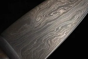 Extreme close-up of Damascus steel blade surface showing characteristic wavy banding pattern of Wootz steel from South India catching light at low angle against dark background